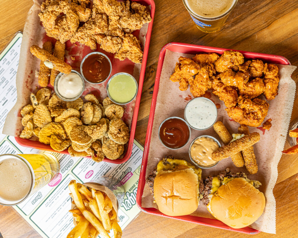 Two colorful red trays loaded with Bareburger's Triple Lindy shareable platters. The trays feature crispy fried chicken, saucy popcorn chicken, golden pickle chips, spicy jalapeño mozzarella sticks, and mini Wagyu Smash Burgers, served with a trio of dipping sauces. Fresh-cut fries and cold draft beers are on the side, creating a crave-worthy, crowd-pleasing table spread.
