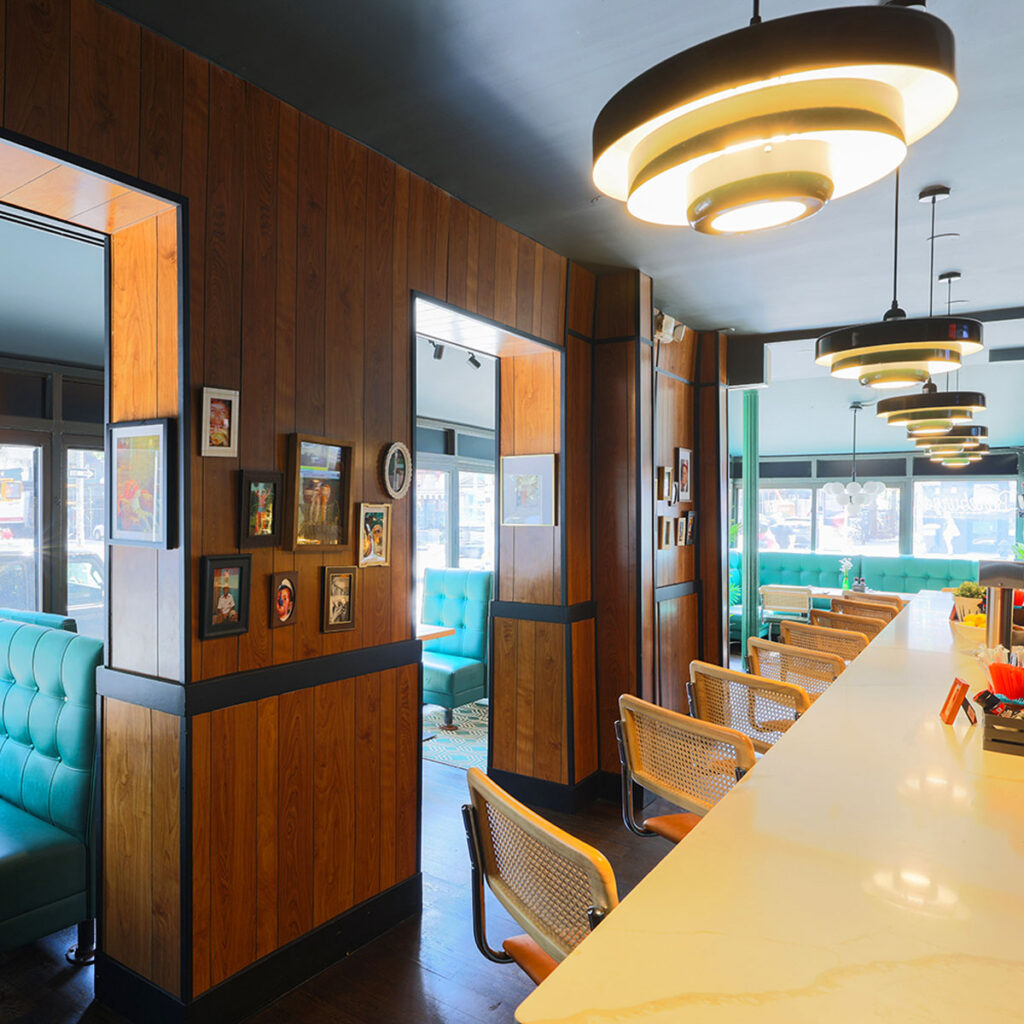Interior of Bareburger Smash Club in Murray Hill featuring retro wood paneling, teal booth seating, mid-century modern lighting, and a marble countertop bar with woven chairs