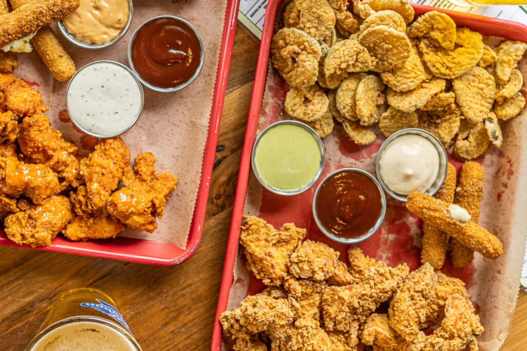 Two colorful red trays loaded with Bareburger's Triple Lindy shareable platters. The trays feature crispy fried chicken, saucy popcorn chicken, golden pickle chips, spicy jalapeño mozzarella sticks, and mini Wagyu Smash Burgers, served with a trio of dipping sauces. Fresh-cut fries and cold draft beers are on the side, creating a crave-worthy, crowd-pleasing table spread.